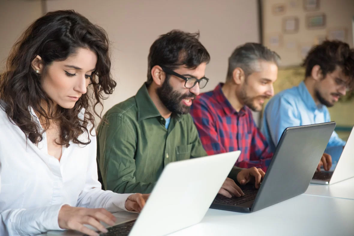 Four people sit side by side at a desk, focused on working on their laptops—perhaps learning how to be a good sourcer in a classroom or office setting, each typing or looking intently at their screen.
