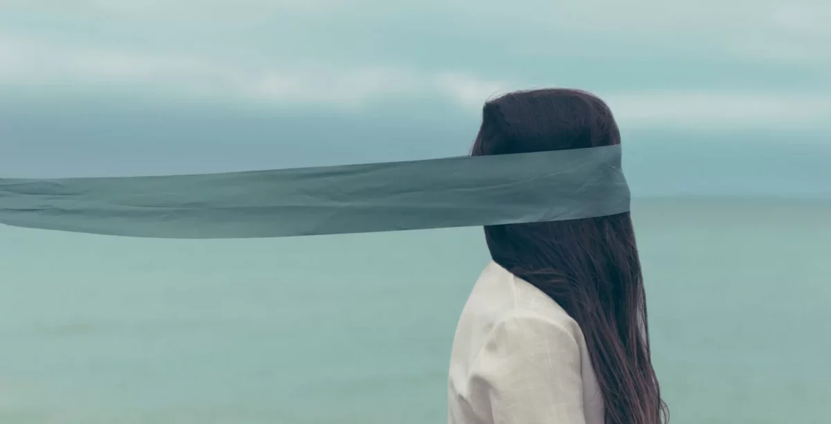 A woman with long brown hair stands by the sea, facing sideways. A sheer blue-gray scarf, like someone working in recruitment advice might wear, covers her eyes and blows behind her in the calm, muted air.